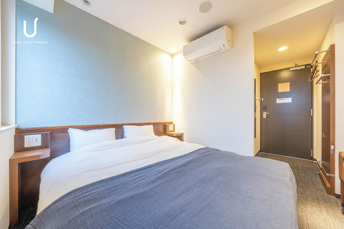 A hotel room with a large bed, a bedside table, an air conditioning unit, and a door leading to a hallway.