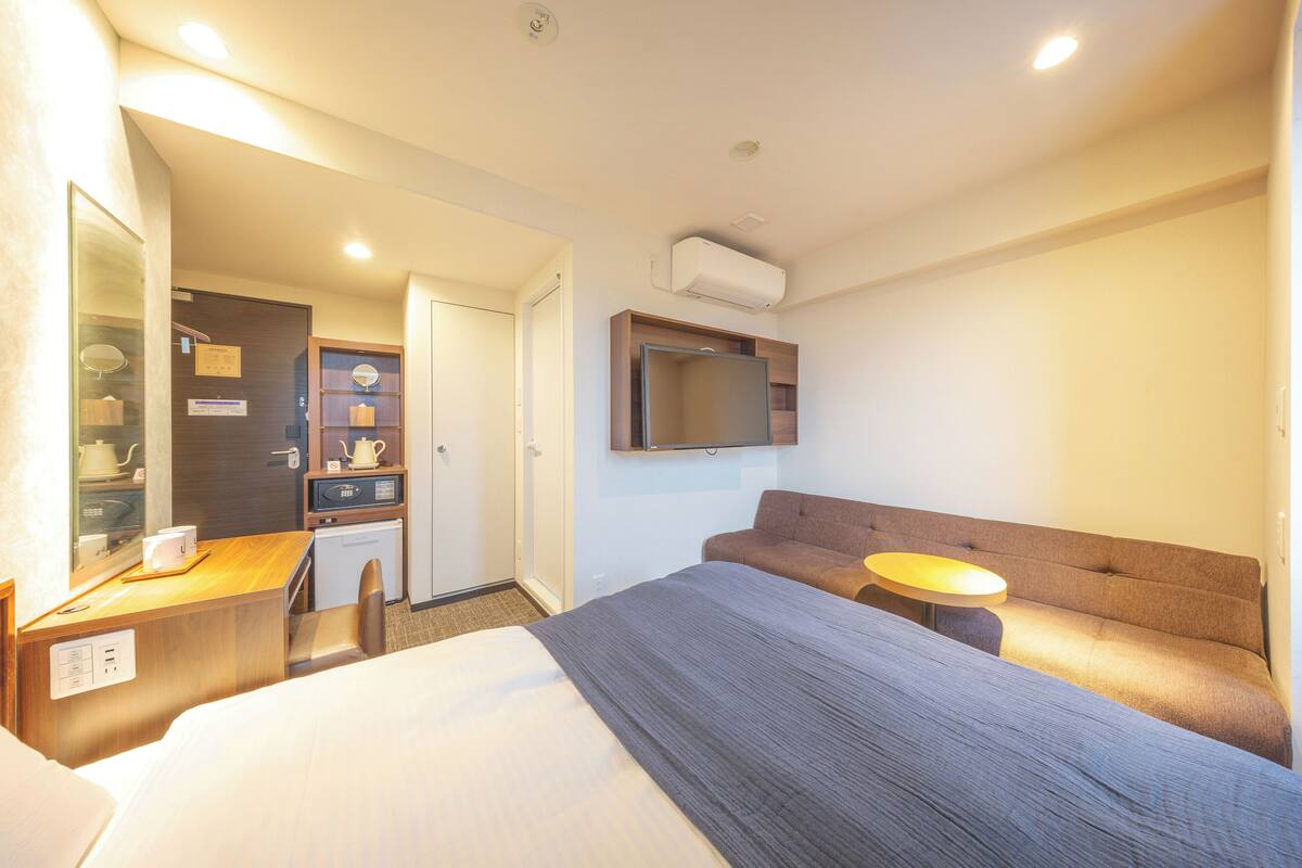 A hotel room with a bed, a desk, a chair, a sofa, a small table, and a TV mounted on the wall.