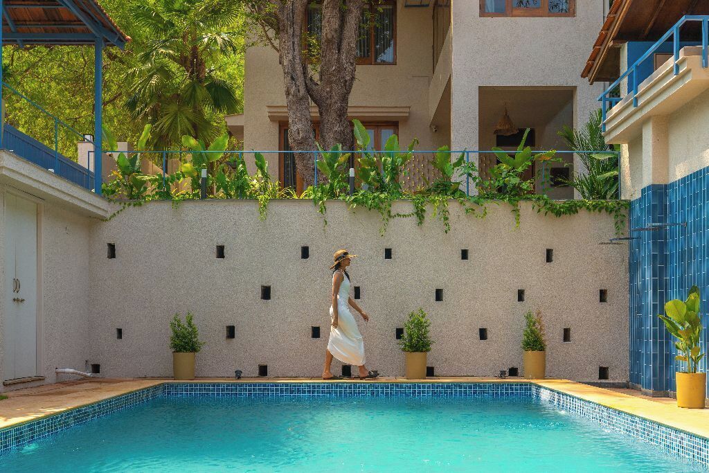 A swimming pool with a tiled edge, surrounded by a low wall with small windows. There are potted plants along the wall, and a tree is visible in the background.