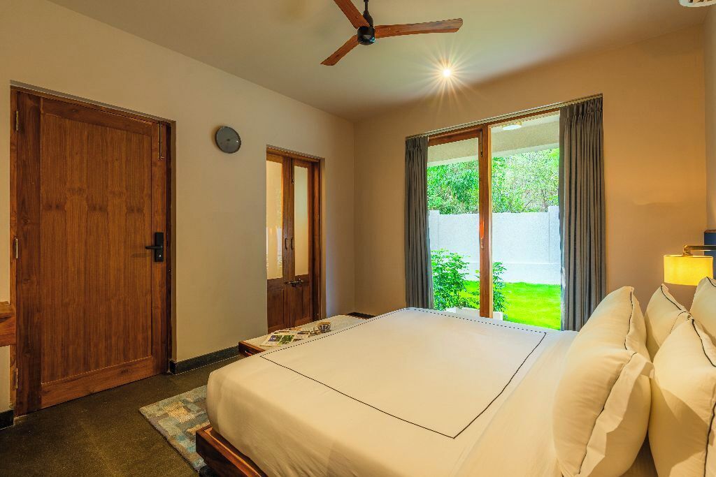 A bedroom with a large bed, wooden doors, a ceiling fan, and a view of greenery outside.