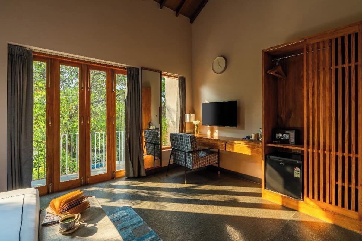 A room with a large sliding glass door leading to a balcony, a flat-screen TV mounted on the wall, a small refrigerator, and a seating area with two chairs and a table.