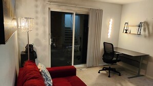 Une pièce avec un canapé rouge, un bureau, une chaise, une lampe et une porte-fenêtre coulissante donnant sur un balcon.