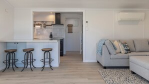 A modern living room with a grey sofa, a small coffee table, and a patterned rug. There are three metal stools with wooden tops. A kitchen area is visible in the background with white cabinets and a black countertop. - Luxury Hideaway at the Lake (Mangakino)