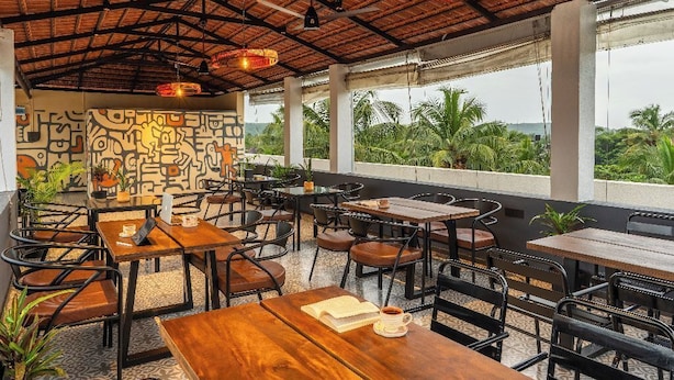 An outdoor cafe with wooden tables and chairs, a patterned wall, and a view of greenery. - The Hosteller Bam Goa Calangute (Calangute)