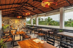 An outdoor cafe with wooden tables and chairs, a patterned wall, and a view of greenery. - The Hosteller Bam Goa Calangute (Calangute)