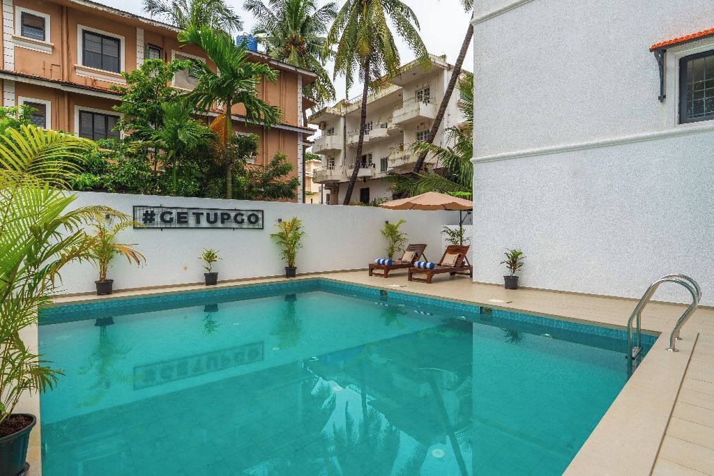 Uma piscina com espreguiçadeiras e plantas em vasos, rodeada por edifícios com varandas e palmeiras.