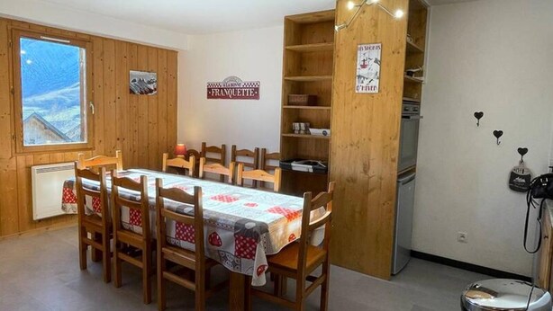 Un comedor con mesa y sillas de madera, vista a las montañas a través de una ventana, y un área de cocina con refrigerador y estufa.