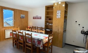 A dining area with a wooden table and chairs, a view of the mountains through a window, and a kitchen area with a refrigerator and stove. - Apartment in La Chal Near Les Sybelles Slopes (Saint-Jean-d'Arves)