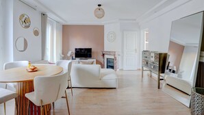 Een moderne woonkamer met een ronde houten tafel, witte stoelen, een witte bank, een flatscreen televisie, een open haard en een grote spiegel.