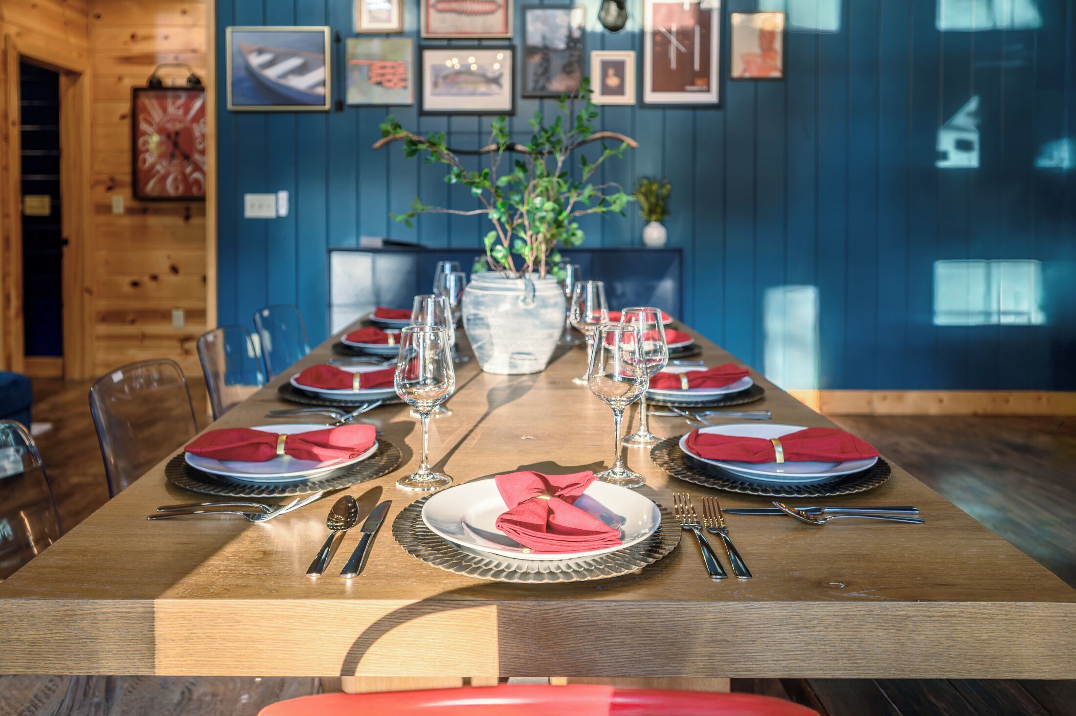 A dining table set with red napkins, wine glasses, and cutlery, with a vase of greenery in the center.