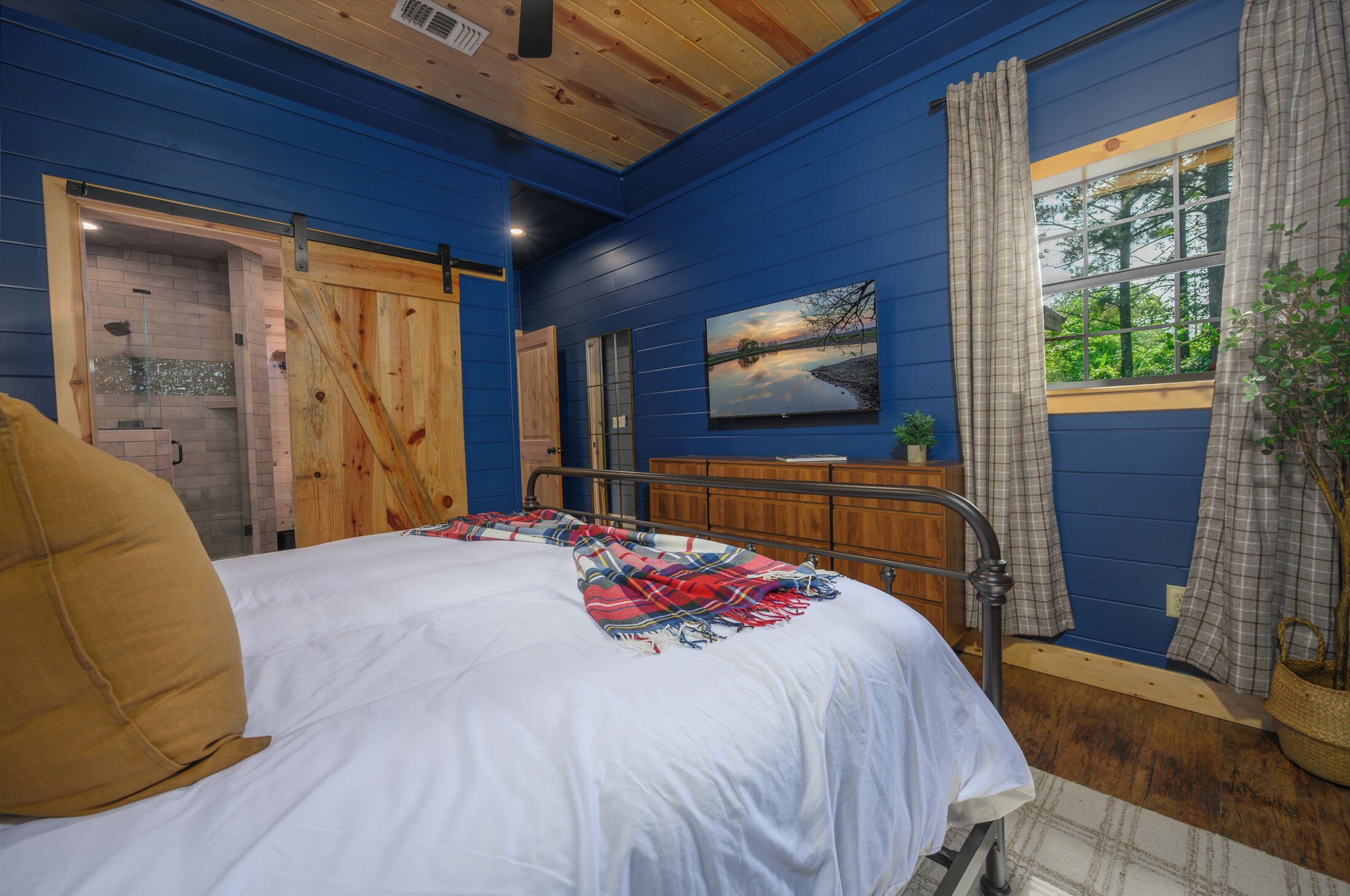 A bedroom with a bed, a wooden door, a window with curtains, and a painting on the wall.