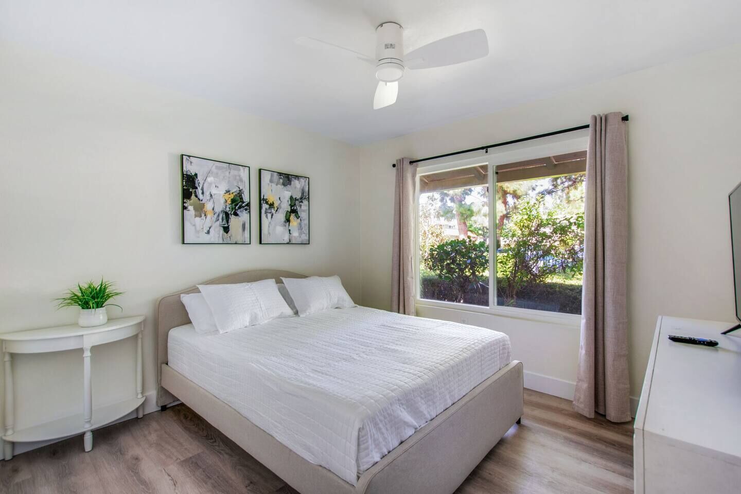 A bedroom with a bed, a nightstand, a plant, and a window with curtains.