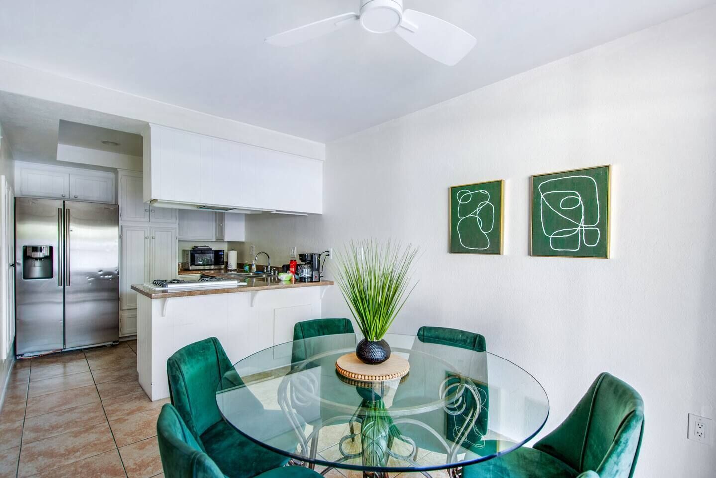 A modern kitchen with white cabinets, a stainless steel refrigerator, and a dining area with a glass table and green chairs.