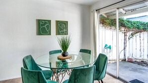 Ein Essbereich mit einem Glastisch, umgeben von grünen Stühlen, zwei abstrakten Gemälden an der Wand und einem Deckenventilator.