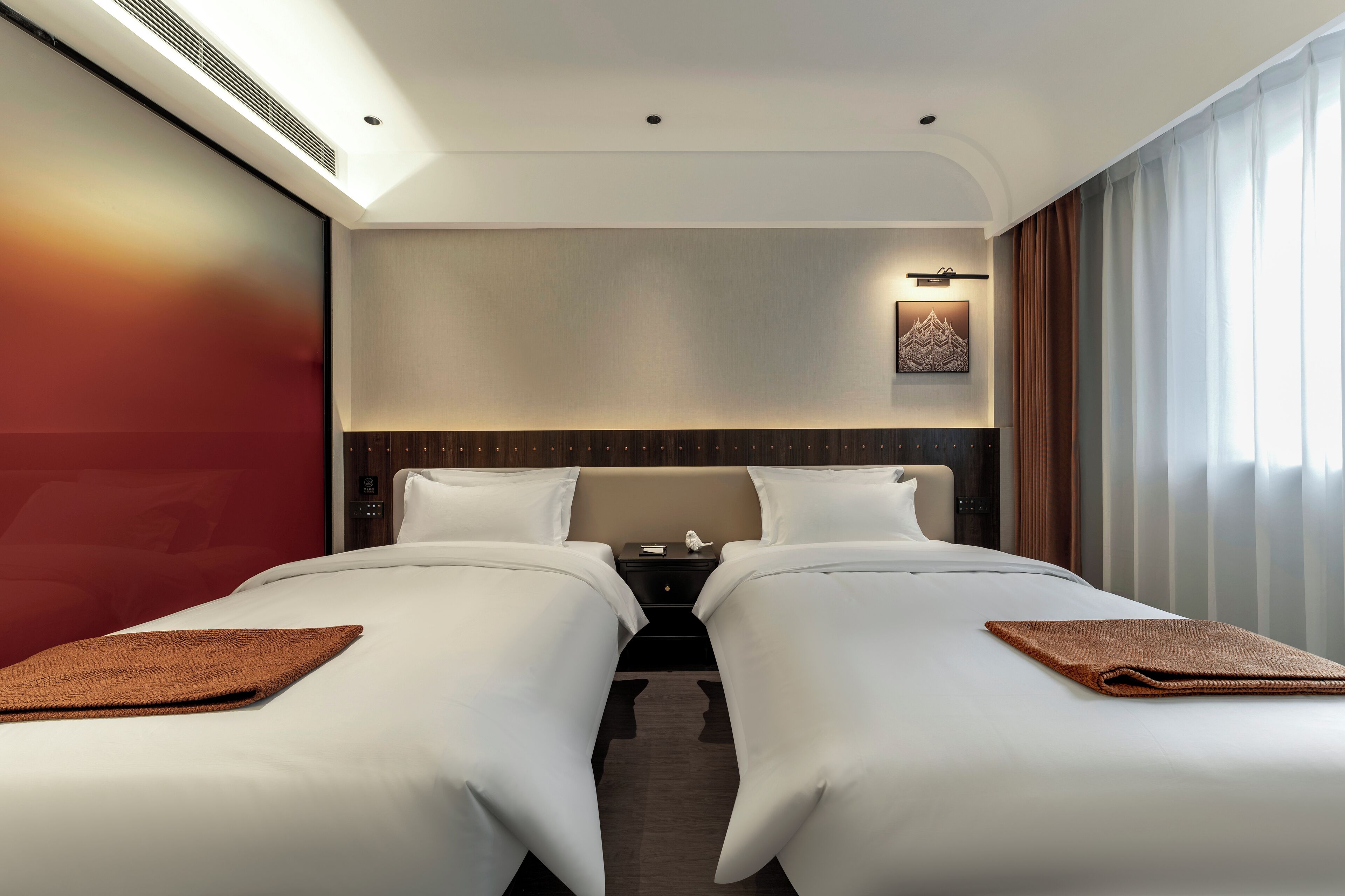 A hotel room with two beds, a nightstand, a wall-mounted light, and a window with curtains.