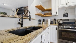 Une cuisine dotée de plans de travail en granit, d’un évier à deux bacs, de meubles blancs, d’étagères en bois et d’une cuisinière en acier inoxydable.
