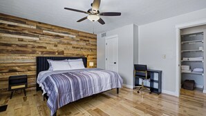 Une chambre avec un mur en bois, un lit recouvert d’une couvre-lit à rayures, un bureau avec une chaise et un ventilateur de plafond.