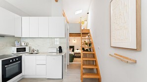 Une cuisine moderne avec des meubles blancs, un four à micro-ondes, un grille-pain et une machine à café. Un escalier en bois mène à une mezzanine aménagée avec un lit et un bureau.