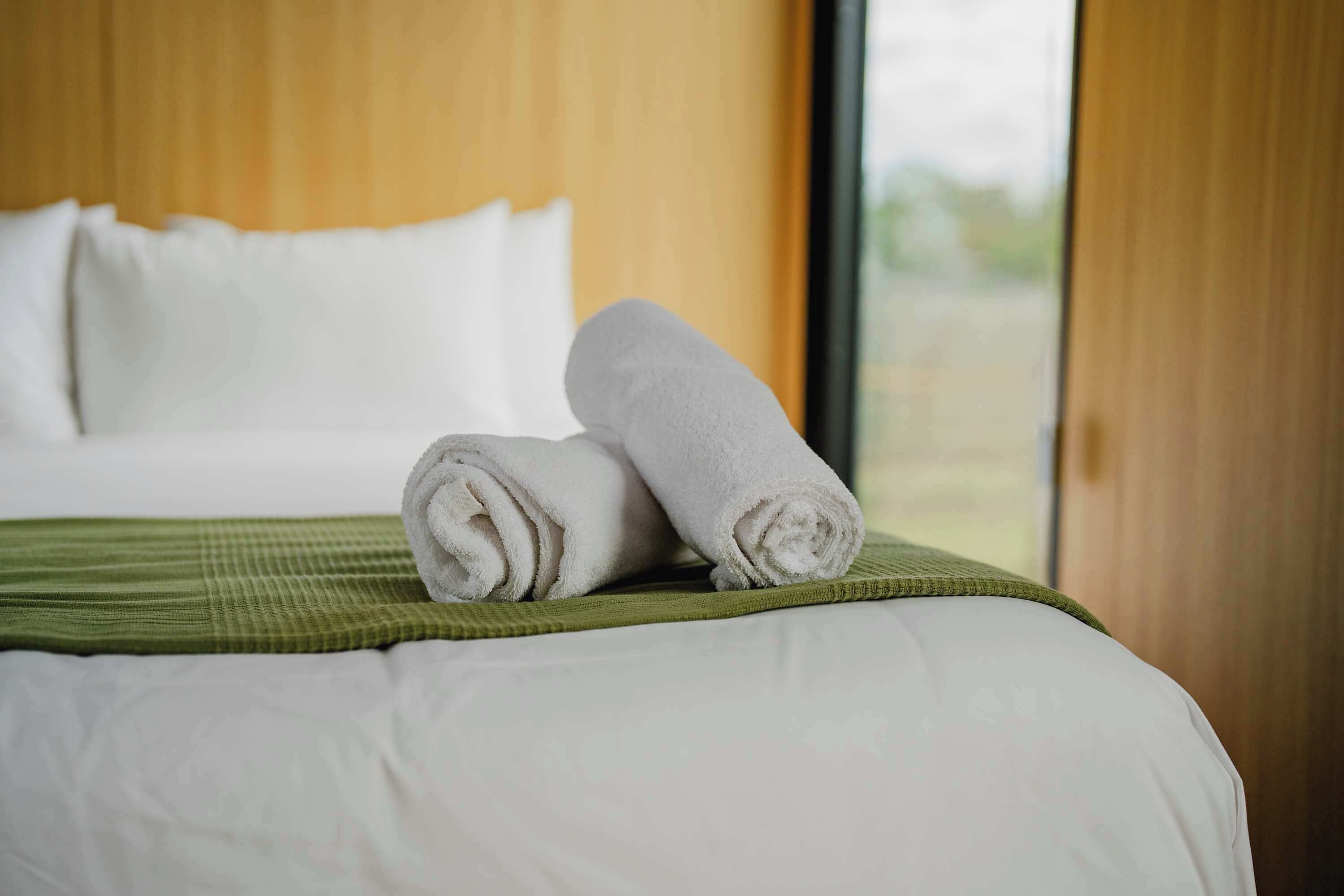 A neatly made bed with white linens and a green bedspread, a window with a view of greenery, and a wooden headboard.