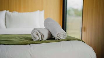 A neatly made bed with white linens and a green bedspread, a window with a view of greenery, and a wooden headboard.