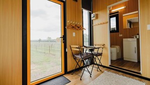 Una habitación moderna con un amplio ventanal corredero que da a una zona ajardinada, una pequeña mesa con sillas, una kitchenette compacta y un espejo sobre el lavamanos.