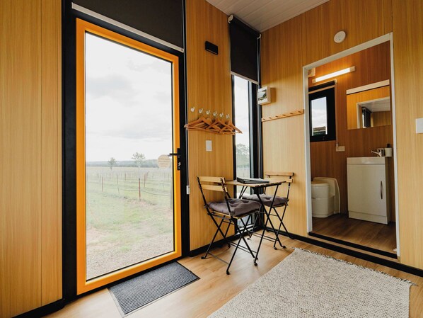 Une chambre moderne dotée d’une grande porte-fenêtre coulissante donnant sur un espace verdoyant, d’une petite table avec des chaises, d’une kitchenette compacte et d’un miroir au-dessus de l’évier.