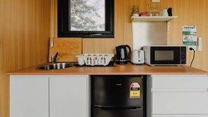 Cocina pequeña con un lavabo, un refrigerador negro, un microondas, un tostador y una tetera. Hay una ventana sobre el lavabo, y una repisa con cristalería y un letrero en la pared.