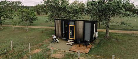 Eine kleine, moderne Holzhütte mit einer Holzterrasse und einer einzigen Tür, umgeben von einem eingezäunten Bereich und Bäumen.