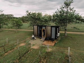 Une petite cabane moderne, dotée d’une terrasse en bois et d’une seule porte, entourée d’une zone délimitée par une clôture et d’arbres.