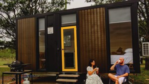 Una cabaña pequeña y moderna con terraza de madera, una barbacoa y un espacio para dormir con cama.