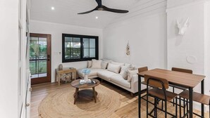 Een moderne woonkamer met een witte bank, een houten salontafel en een eettafel met stoelen. Er is een plafondventilator, een raam met jaloezieën en een decoratief wandkunstwerk.