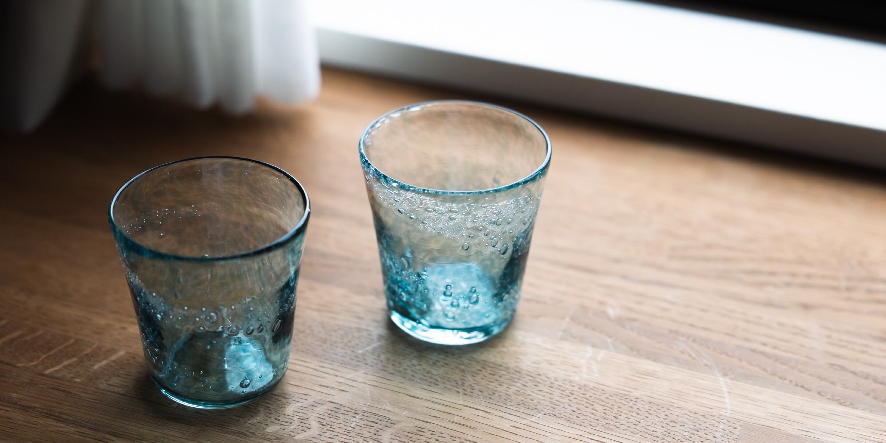 Deux verres d’eau sur une surface en bois, près d’une fenêtre.