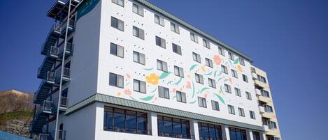 A multi-story hotel with a white facade and colorful floral artwork. It features balconies on the upper floors and a visible fire escape.