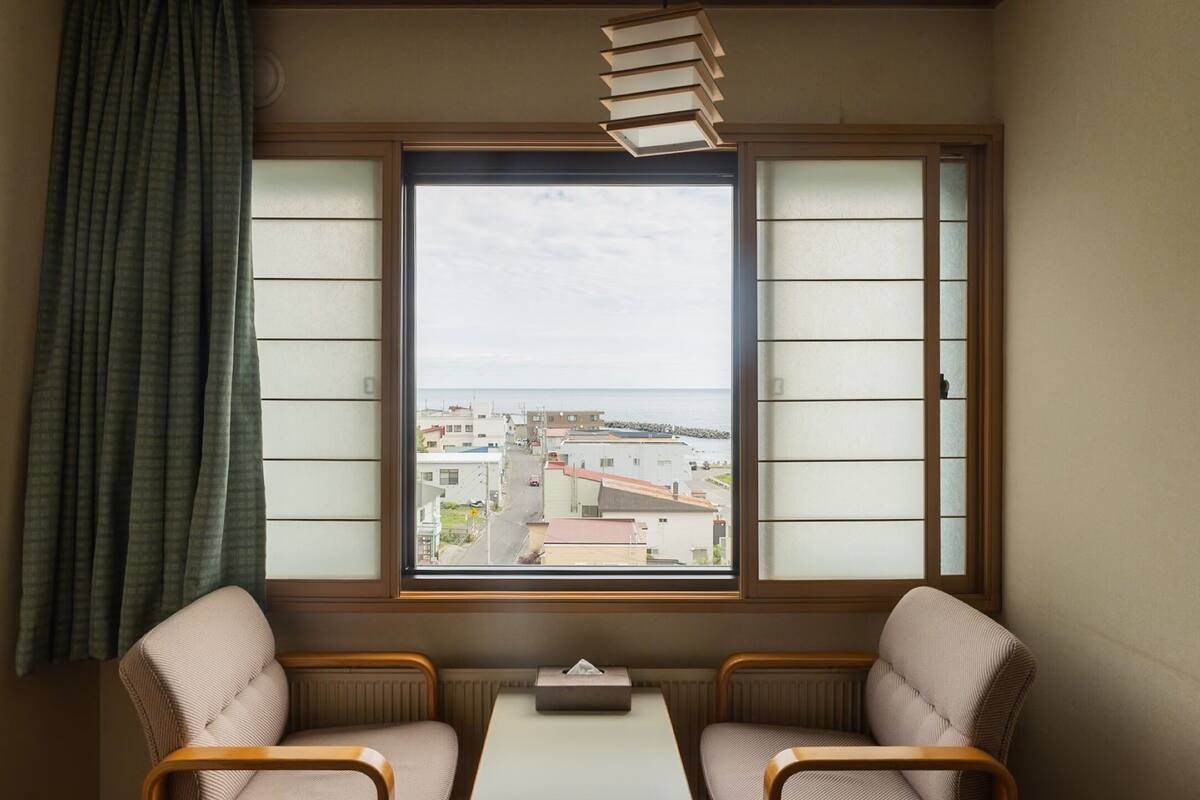 A room with a view of a cityscape through a window with shoji screens, two chairs, a small table, and a hanging light fixture.