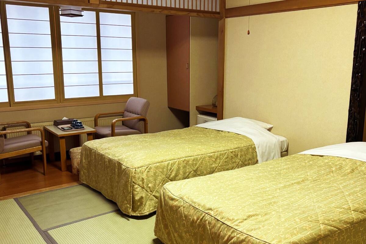 A hotel room with two beds, a chair, a small table, and a window with blinds.