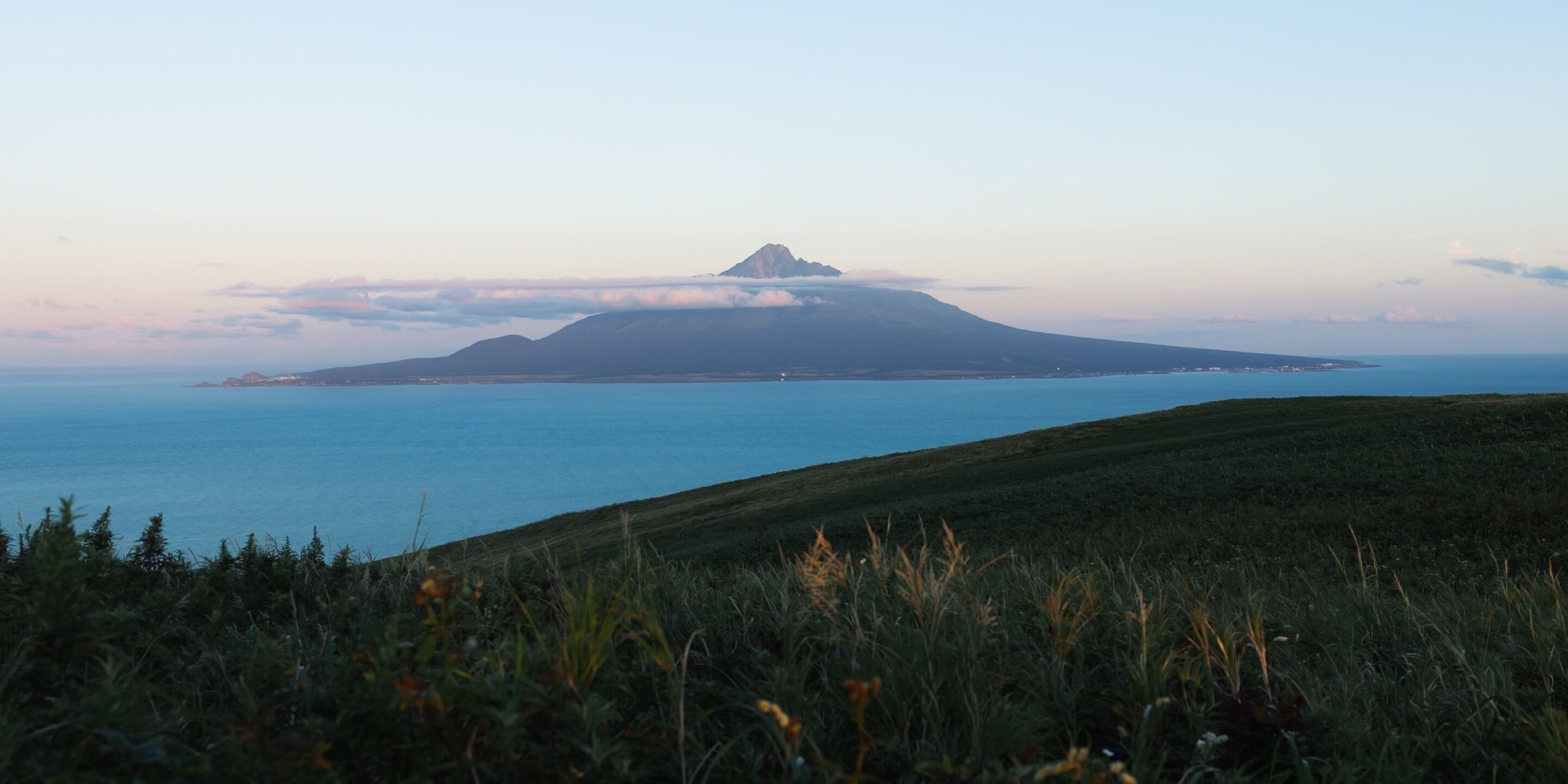 Un paisaje costero con una montaña destacada al fondo y un primer plano cubierto de hierba.