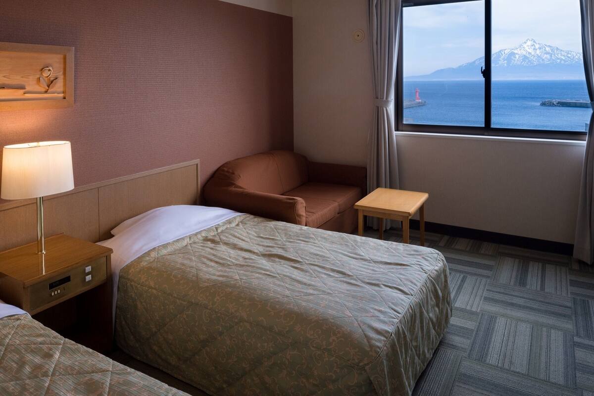 A hotel room with two beds, a small table, a chair, a lamp, and a window with a view of the sea and mountains.
