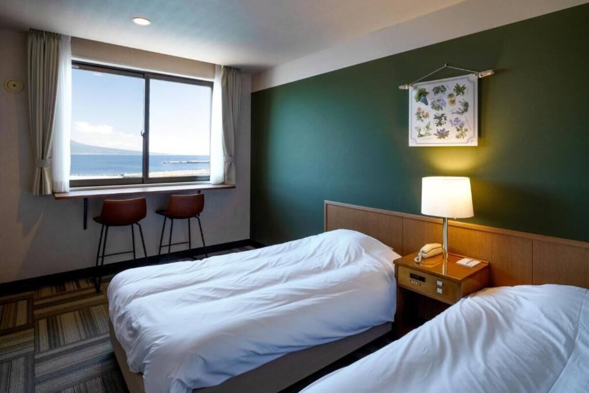 A hotel room with two beds, a window with a view of the sea, a wall-mounted lamp, and a framed floral artwork.