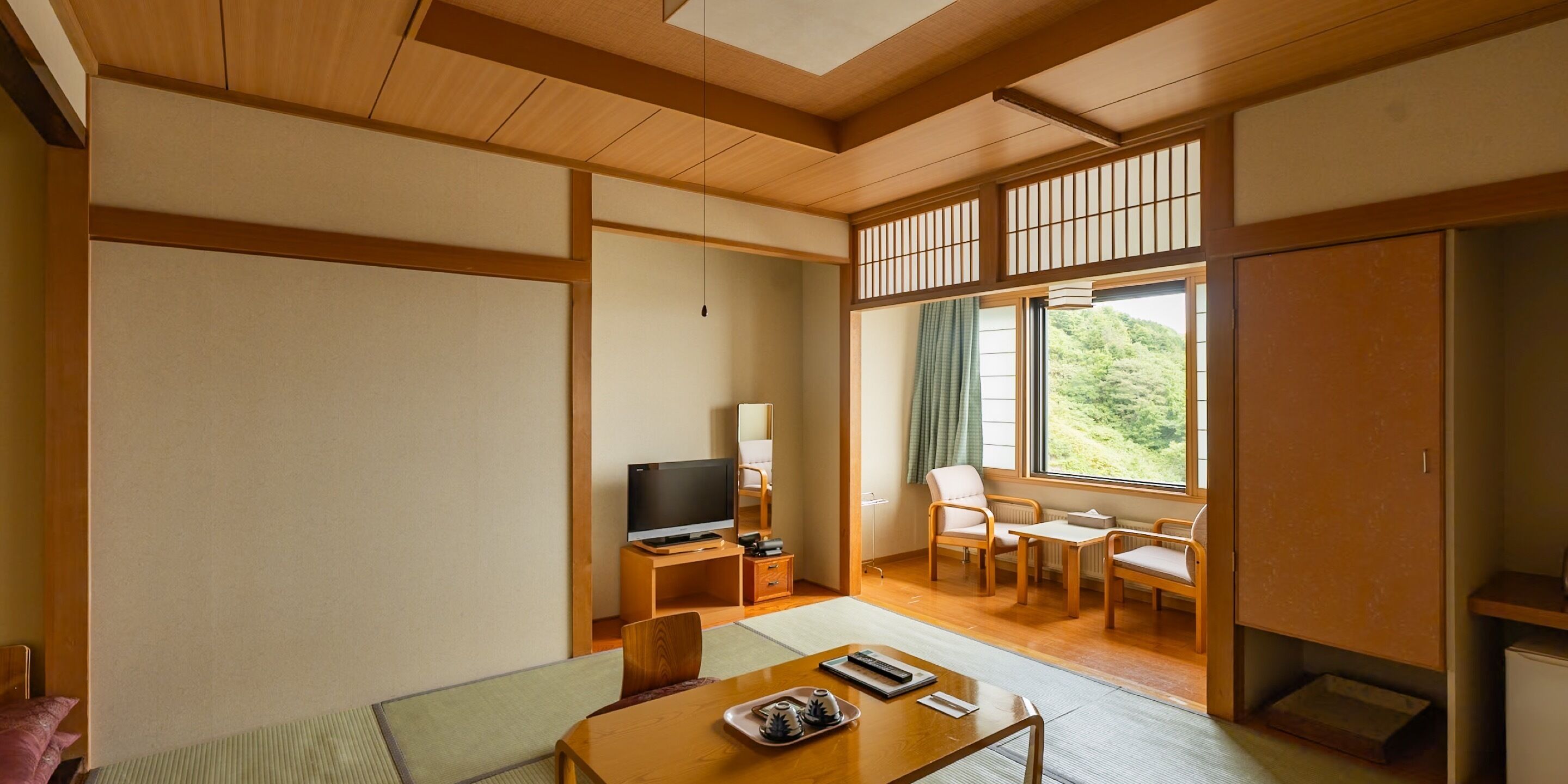 A traditional Japanese-style room with tatami flooring, a television, a desk, and a view of a forest through a sliding door.