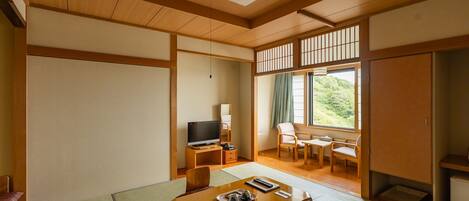 A traditional Japanese-style room with tatami flooring, a television, a desk, and a view of a forest through a sliding door.