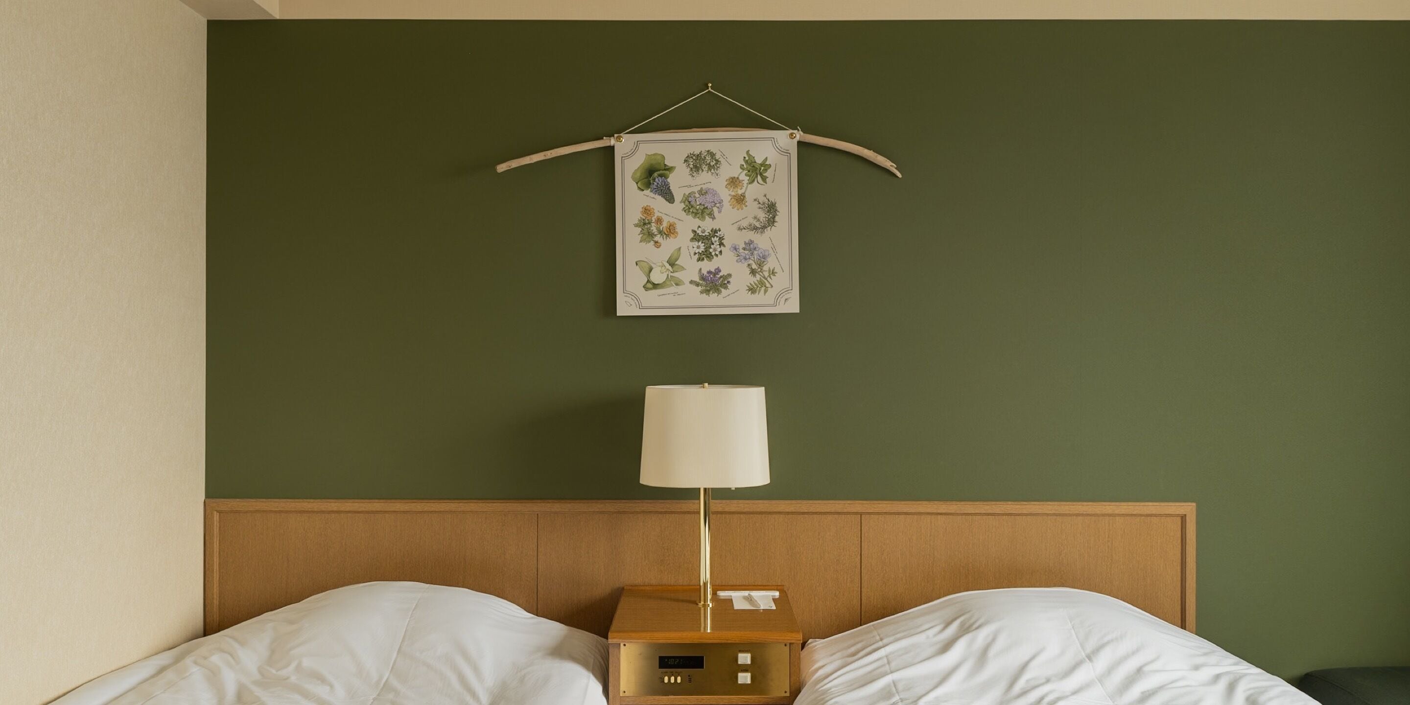 Habitación de hotel con cabecera de madera, cama con ropa blanca, mesita de noche con lámpara e impresión botánica enmarcada en la pared.