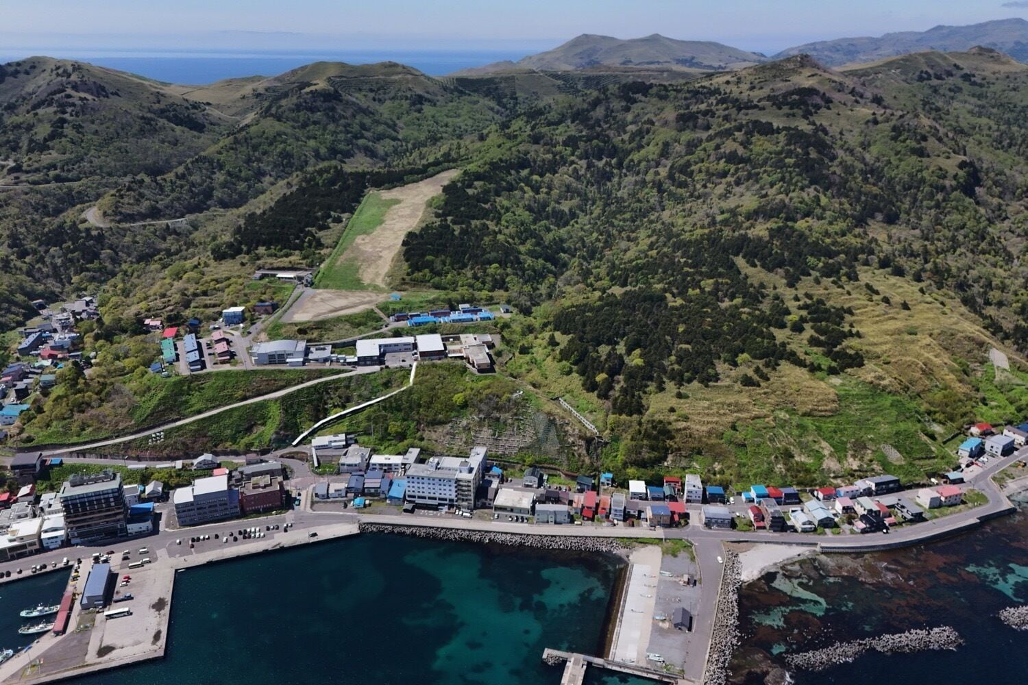 An aerial view of a coastal town with buildings, a marina, and surrounding hills.
