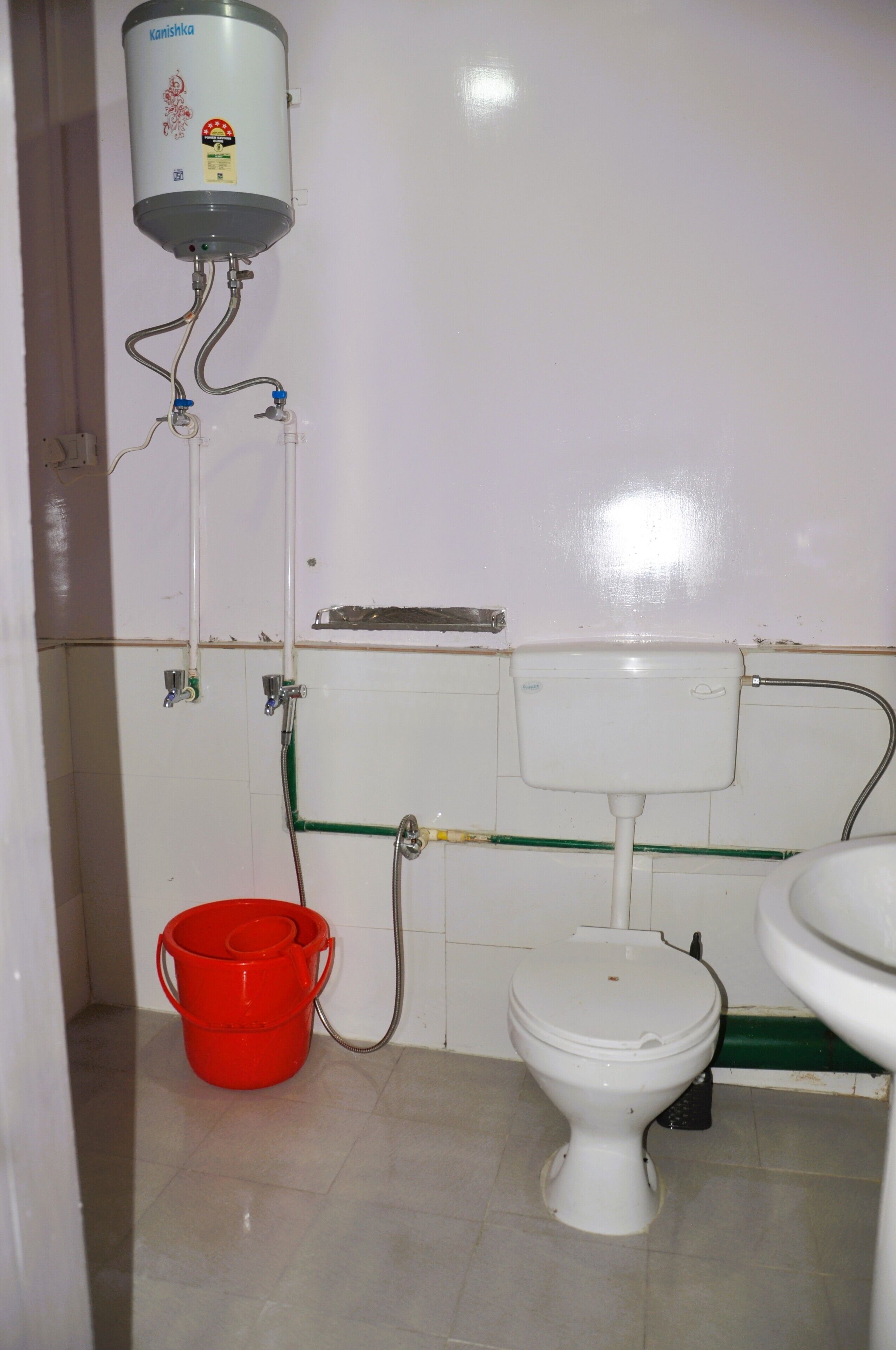 A bathroom with a toilet, a red bucket, a water heater, and a green pipe.