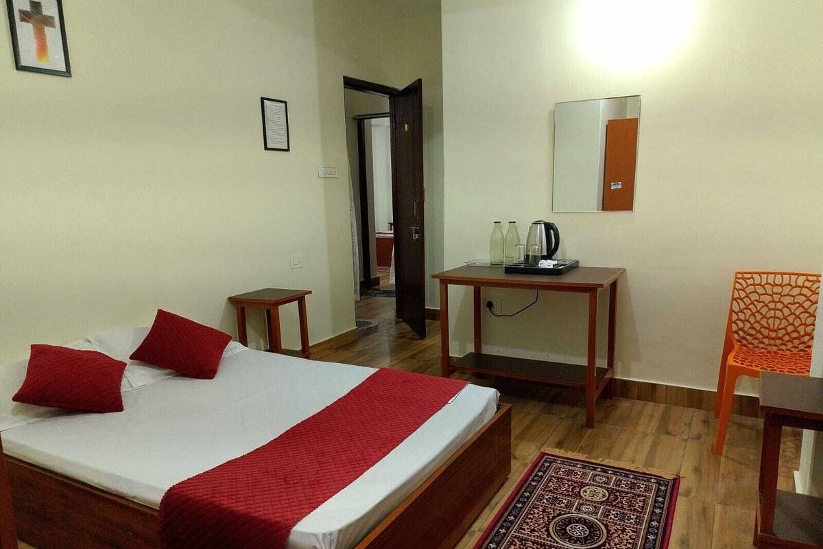 A hotel room with a bed, a small table with a kettle, a chair, and a rug.