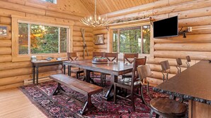 A log cabin interior with a large dining table, benches, and chairs. There are windows with views of trees, a chandelier, and a flat-screen TV mounted on the wall. - Big Sky Cozy Cabin | Lone Peak Views + Hot Tub (Gallatin Gateway)