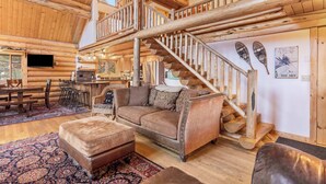 A log cabin interior with a wooden staircase, a sofa, and a dining area. - Big Sky Cozy Cabin | Lone Peak Views + Hot Tub (Gallatin Gateway)