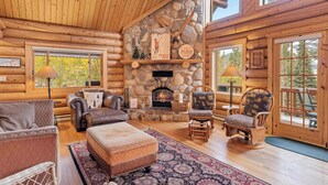 A log cabin interior with a stone fireplace, leather furniture, and large windows. - Big Sky Cozy Cabin | Lone Peak Views + Hot Tub (Gallatin Gateway)