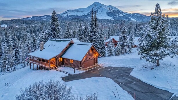 A log cabin with a snow-covered roof, surrounded by snow-laden trees and a mountain in the background. - Big Sky Cozy Cabin | Lone Peak Views + Hot Tub (Gallatin Gateway)