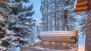 A hot tub in a snowy outdoor setting with a stone wall and wooden cabin in the background. - Big Sky Cozy Cabin | Lone Peak Views + Hot Tub (Gallatin Gateway)