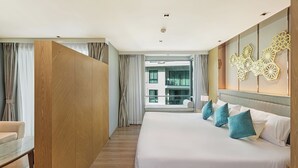 A modern hotel room with a large bed, a wooden wardrobe, a seating area, and a view of a building through the window. - Studio in LaVita Condo in Rawai 7501 (Rawai)